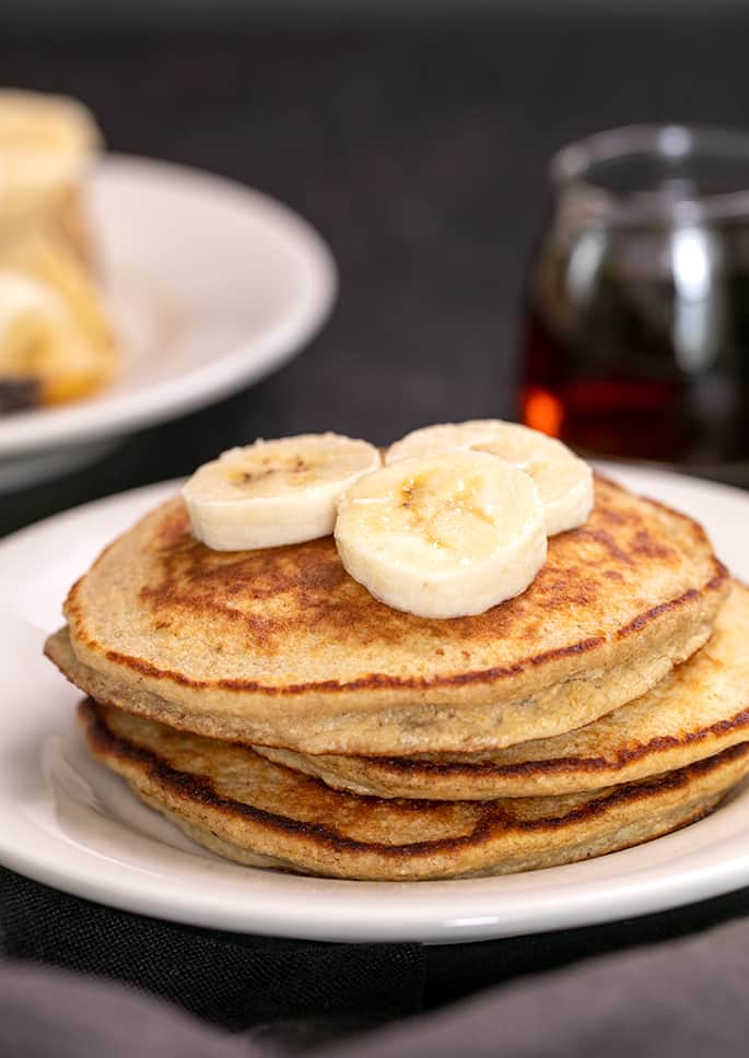 Stack of 3 banana pancakes on white plate with three fresh banana slices on top