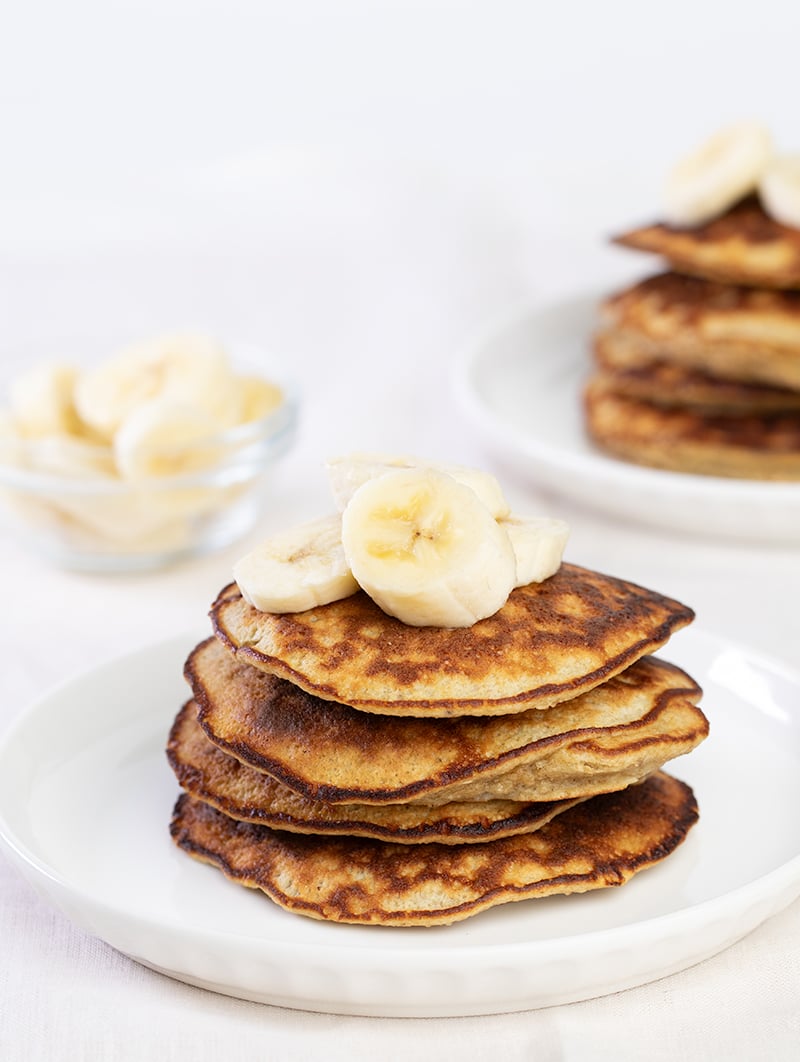 Stack of 4 banana pancakes with banana slices on top on white plate