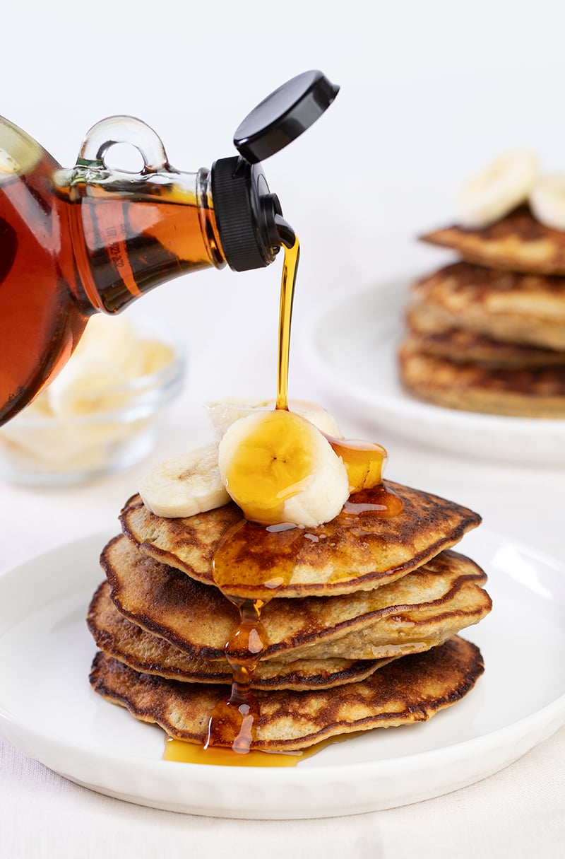 Syrup pouring from bottle onto stack of 4 banana pancakes with banana slices