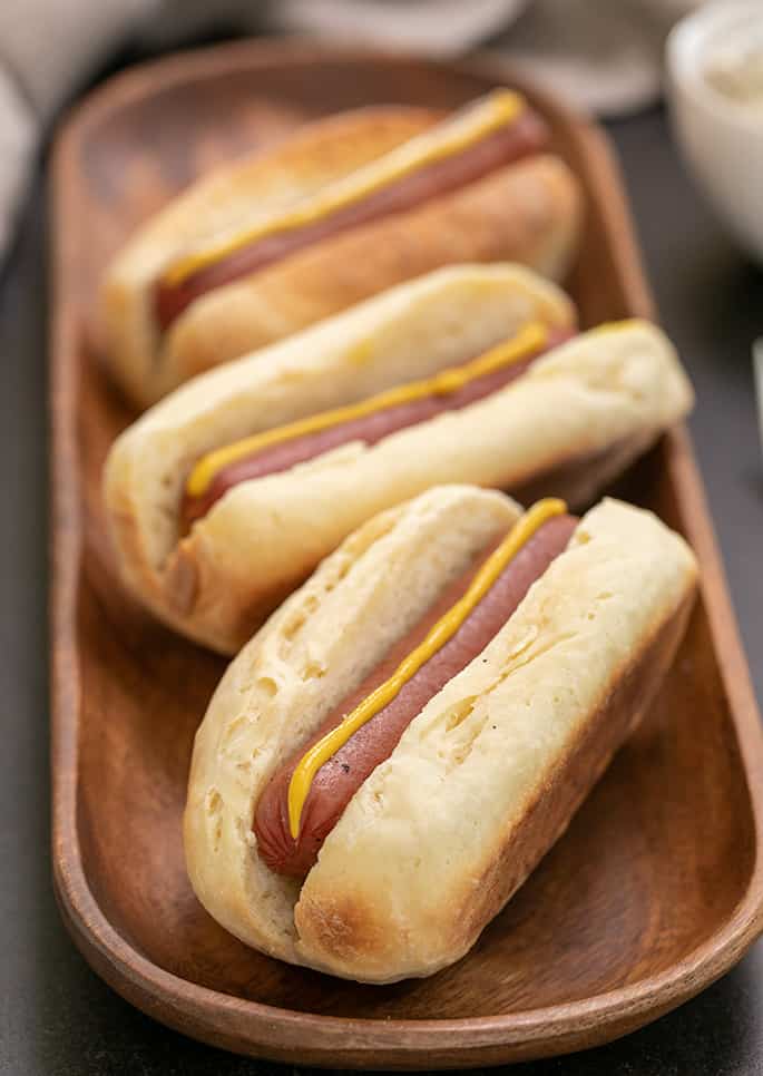 3 hot dogs in buns with mustard on long rectangular wooden tray