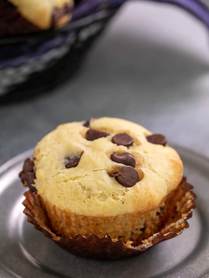 Chocolate chip muffin with muffin liner partially peeled off on a small pewter plate and muffin basket in background