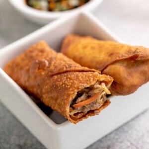 2 gluten free egg rolls in egg roll wrappers in small square white dish with bite taken from one