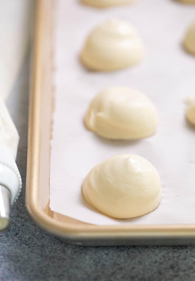 Raw mounds of choux pastry on white paper on baking sheet with piping bag