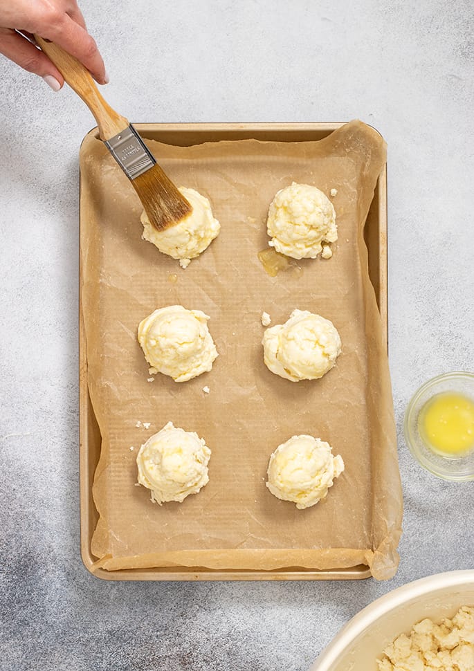 Hand holding pastry brush brushing melted butter on one of 6 raw biscuits on tray lined with brown paper