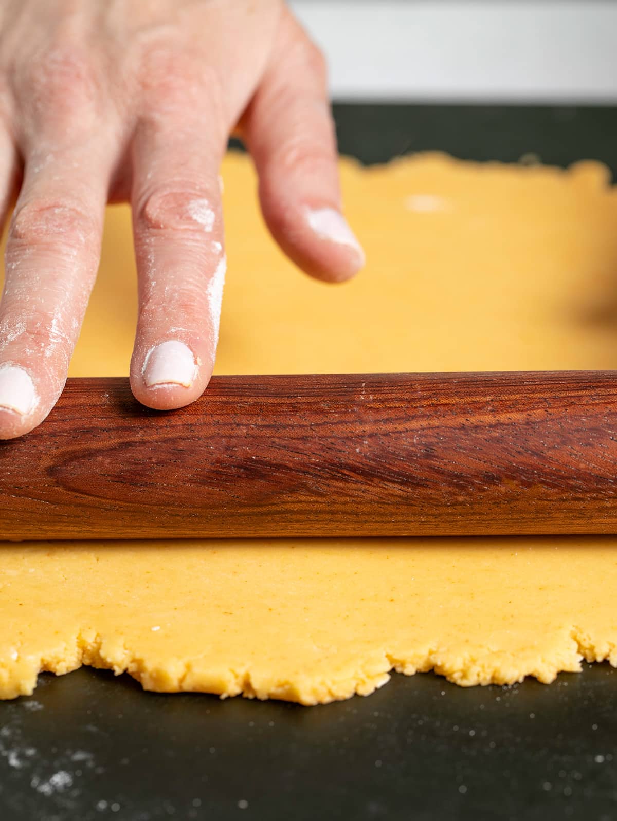 Hands rolling a brown rolling pin over light orange dough on black surface