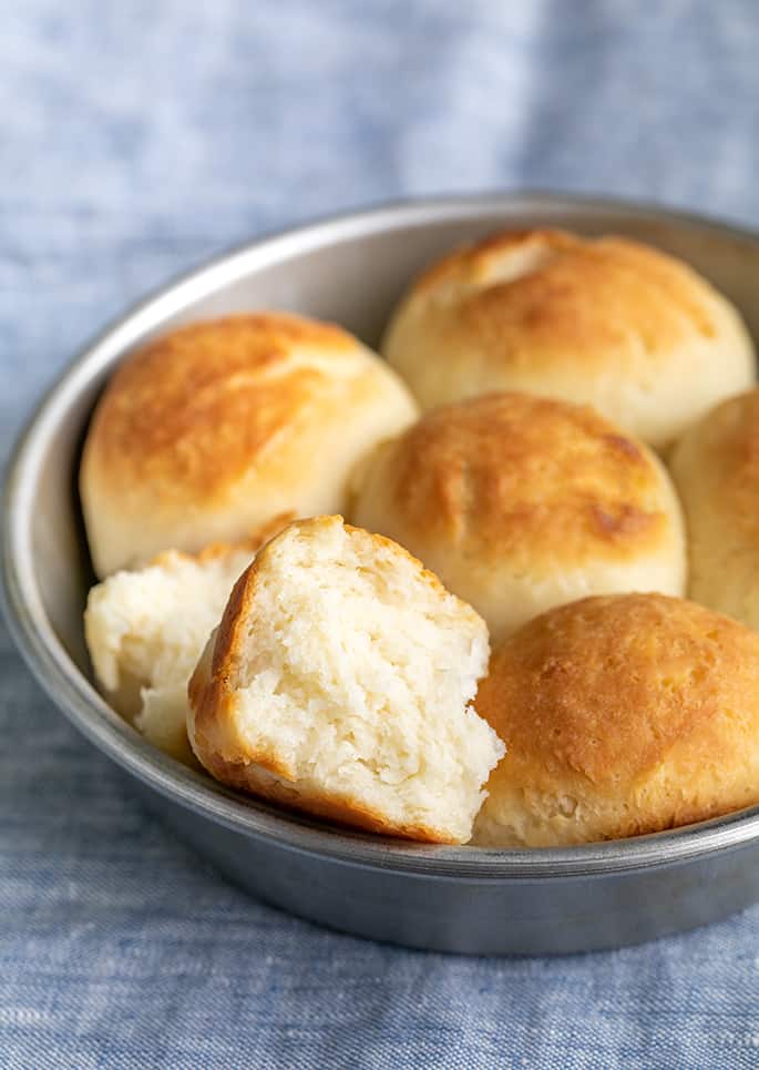 round metal tin with 6 gluten free dinner rolls on blue cloth, one roll broken in half