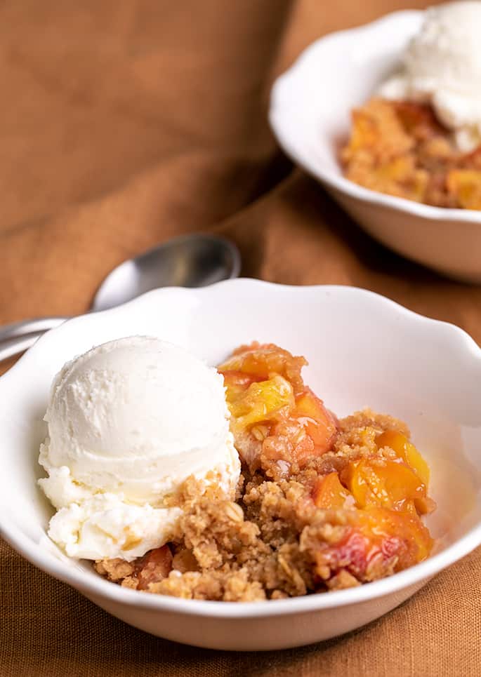 Crispy crumble topping and cooked peach filling with scoop of vanilla ice cream in white bowl on brown cloth