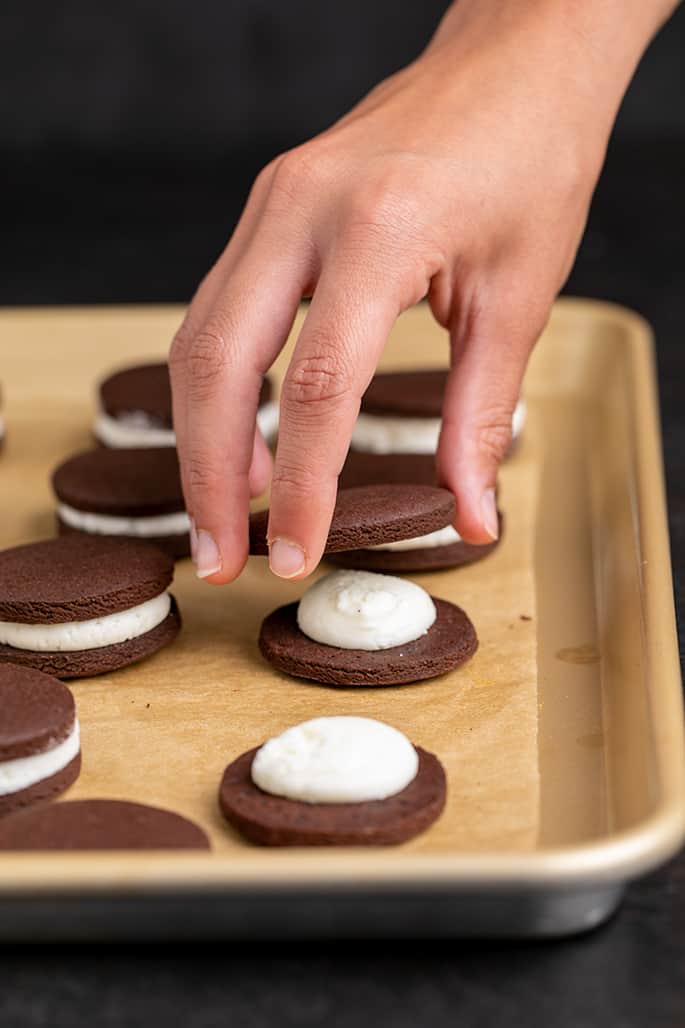 Hand holding a baked round chocolate cookie and placing it on top of filling that's on another cookie