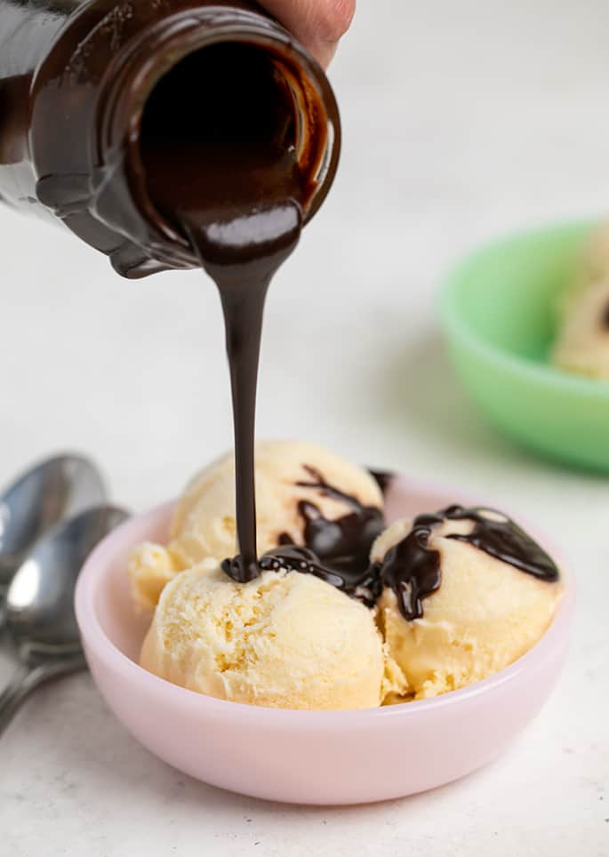 Hot fudge sauce pouring from a small glass jar onto vanilla ice cream scoops in small pink bowl