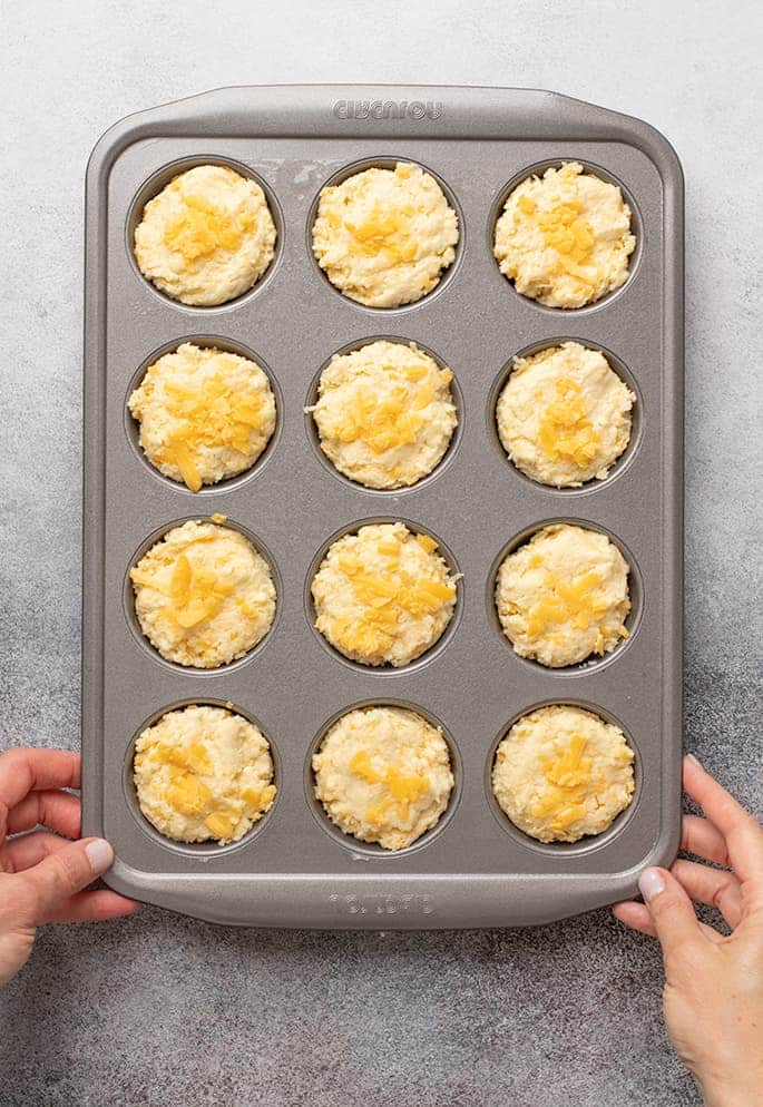 Overhead image of hands in corners of muffin tin with raw yellow batter in wells