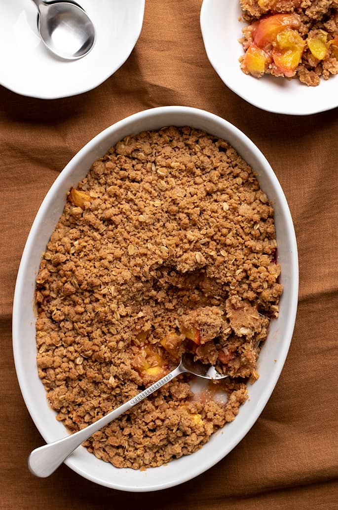 Overhead image of peach crisp in white oval serving dish with serving spoon and two small white bowls one with a serving