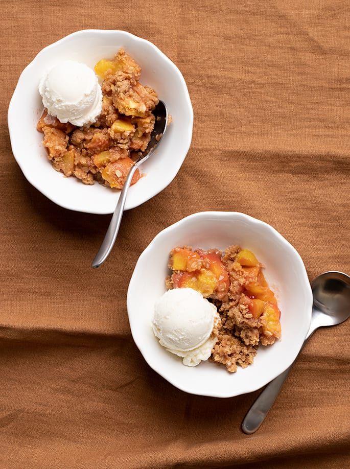 Overhead image of gluten free peach crisp with one scoop of ice cream and a spoon in two small white bowls on brown cloth