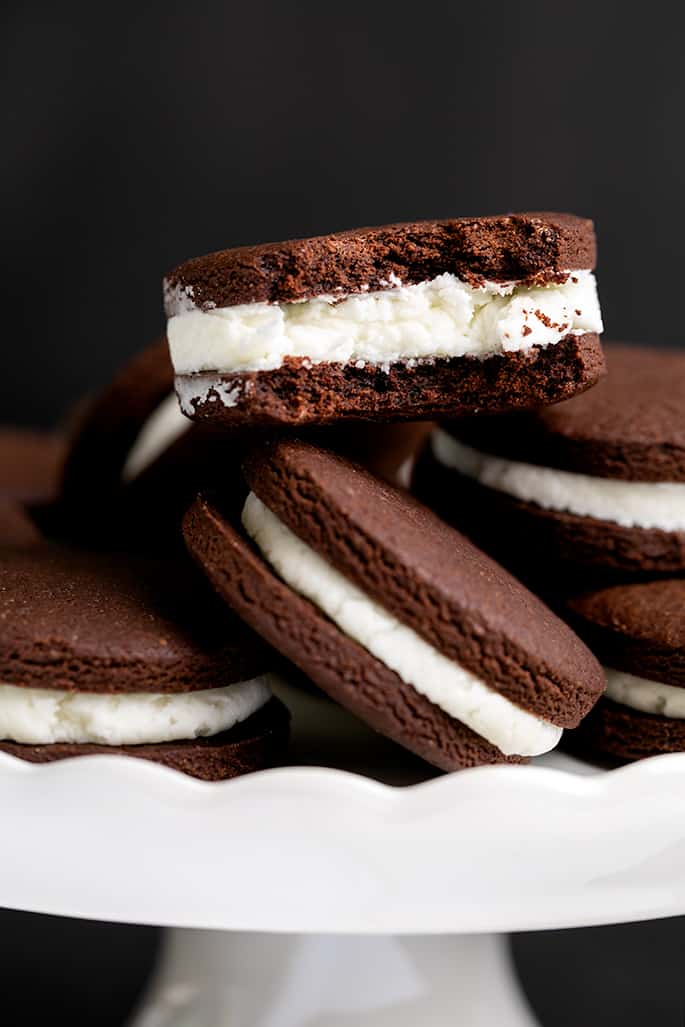 Pile of chocolate sandwich cookies with white filling on a white scalloped cake plate with one cookie on top with a bite taken