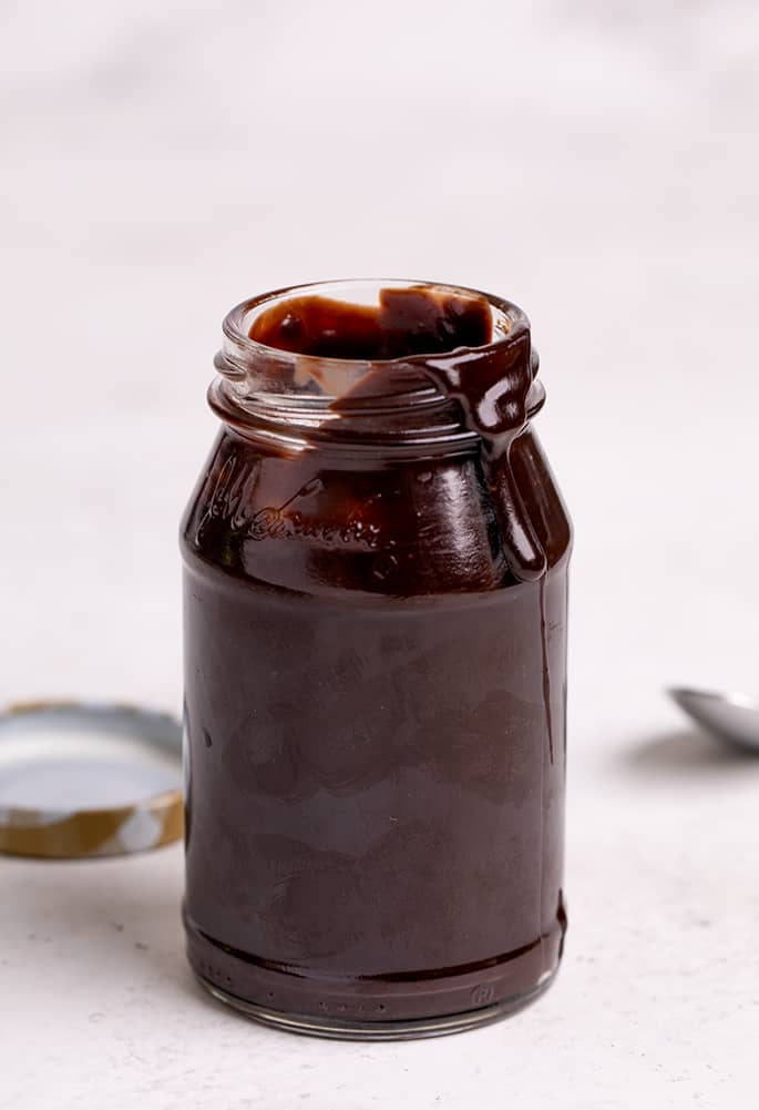 Small glass jar filled with brown hot fudge sauce with some dripping down the side