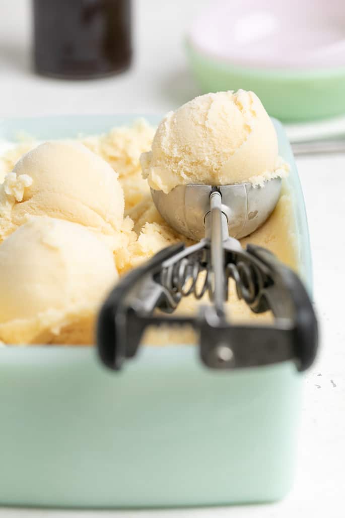 Vanilla ice cream in a spring-loaded ice cream scoop resting in a green rectangular freezer container with more ice cream