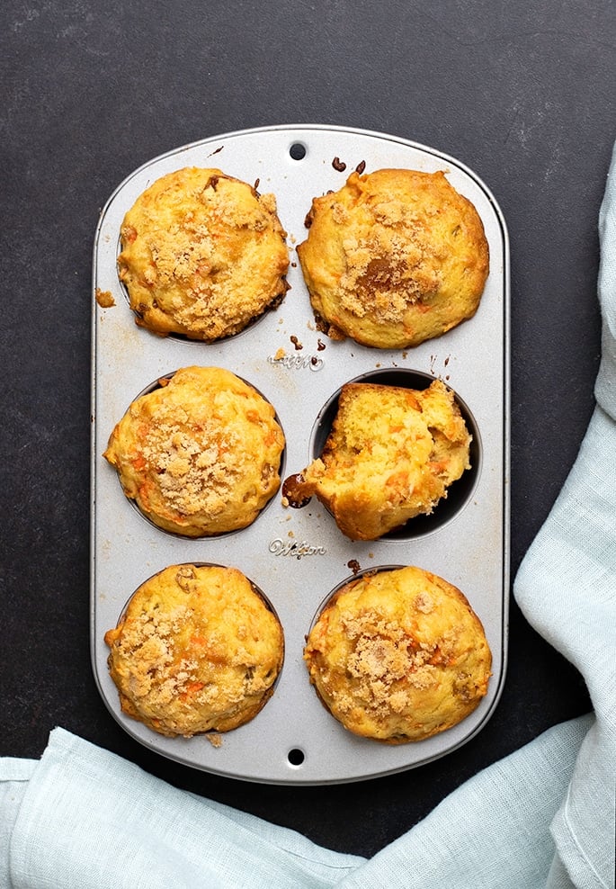 Overhead image of 6 carrot muffins in a muffin tin with one broken in half and a light blue cloth