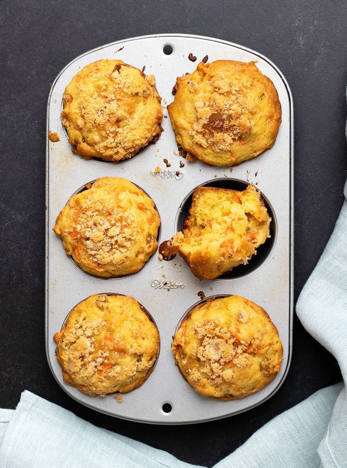 Overhead image of 6 carrot muffins in a muffin tin with one broken in half and a light blue cloth