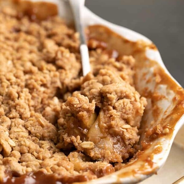 Serving spoon with serving of baked apple crisp sitting in white baking dish on metal tray