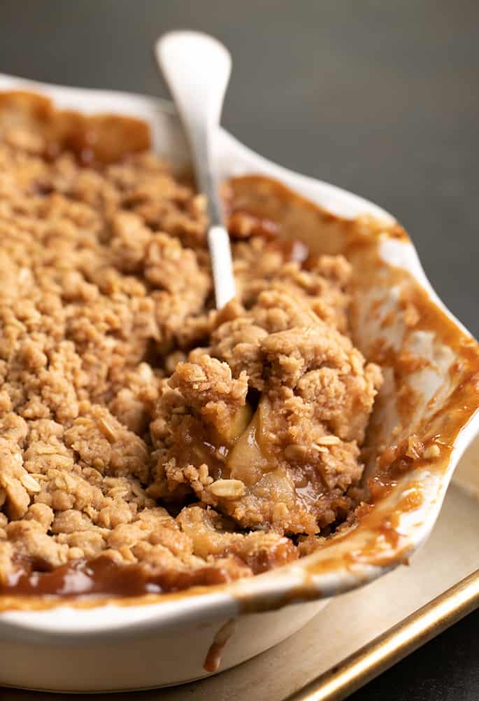 Serving spoon with serving of baked apple crisp sitting in white baking dish on metal tray