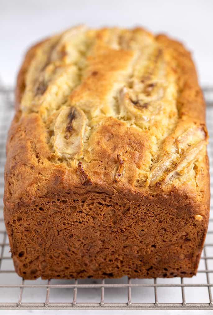 Whole baked brown quick bread with baked banana slices on top on wire rack on white surface