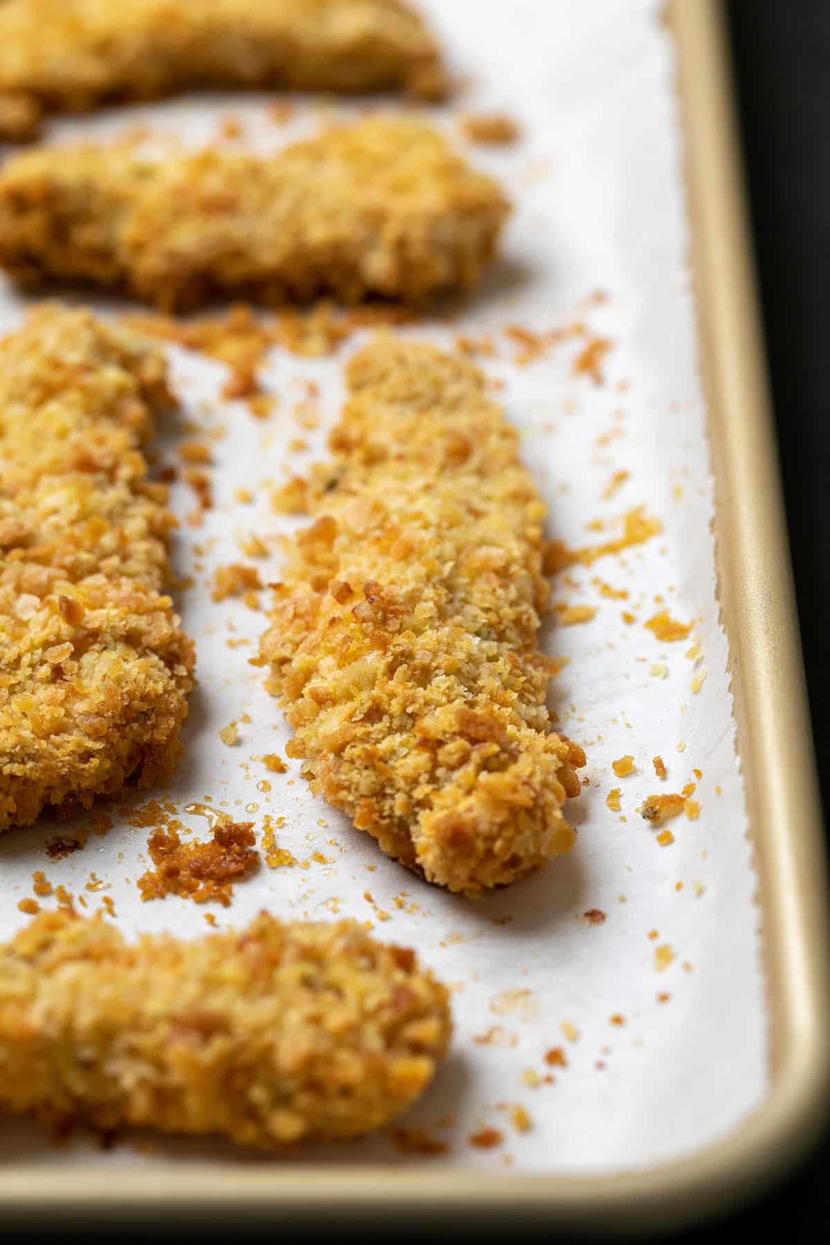 Baked breadcrumb-crusted chicken strips on white paper in corner of gold baking sheet