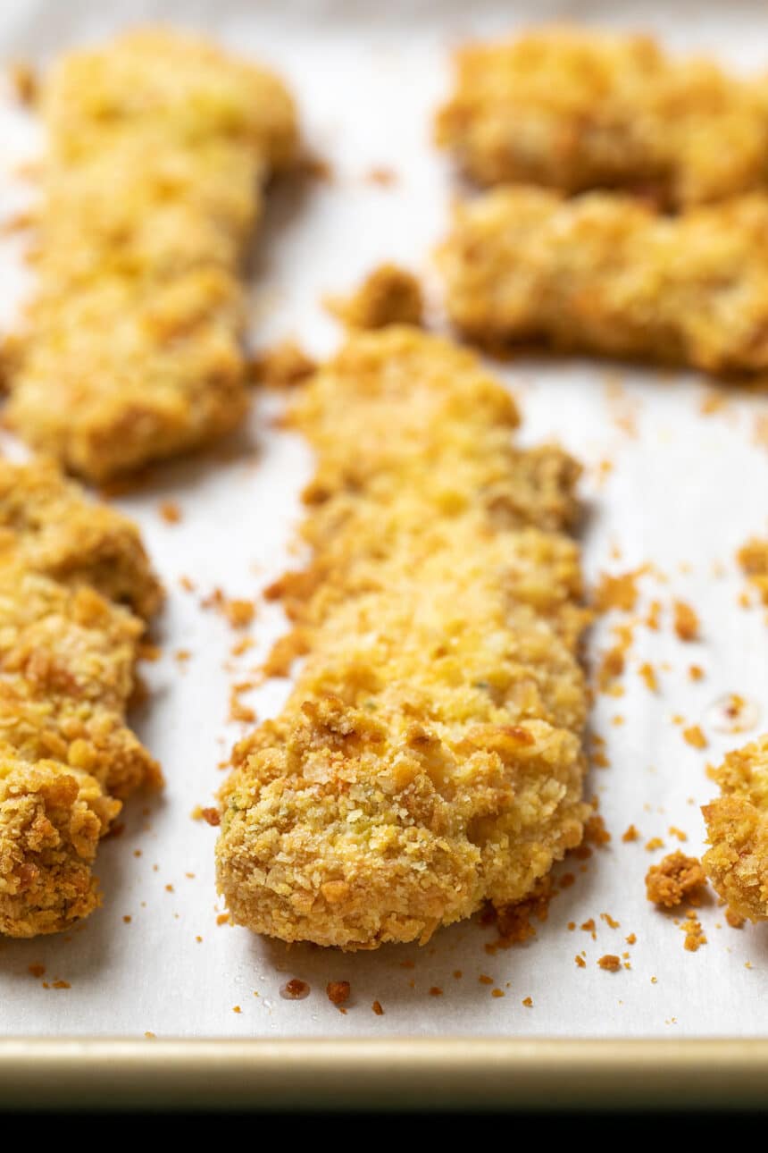 Baked crispy chicken strips with breadcrumb coating on white paper on gold rimmed baking sheet