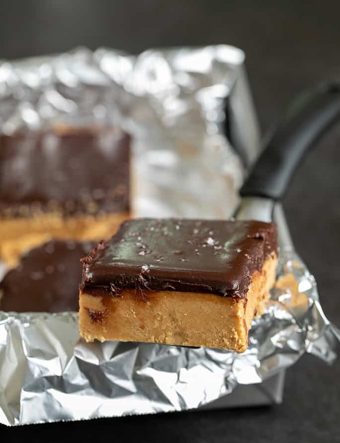 Chocolate topped peanut butter bar on metal spatula on side of foil wrapping on square metal pan