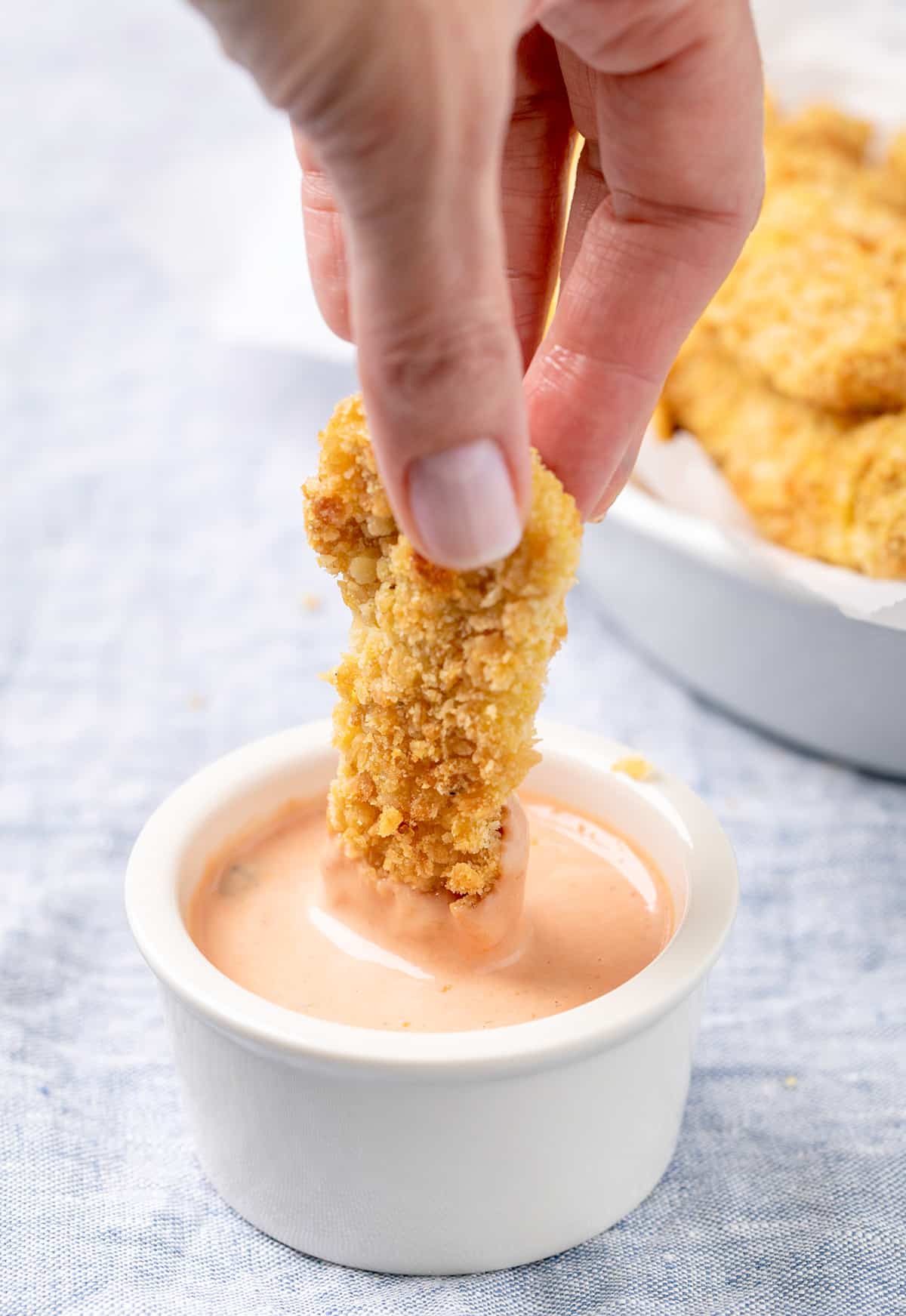 Fingers dipping baked chicken tender strip in orange sauce in small white ramekin on blue cloth with more chicken strips in background