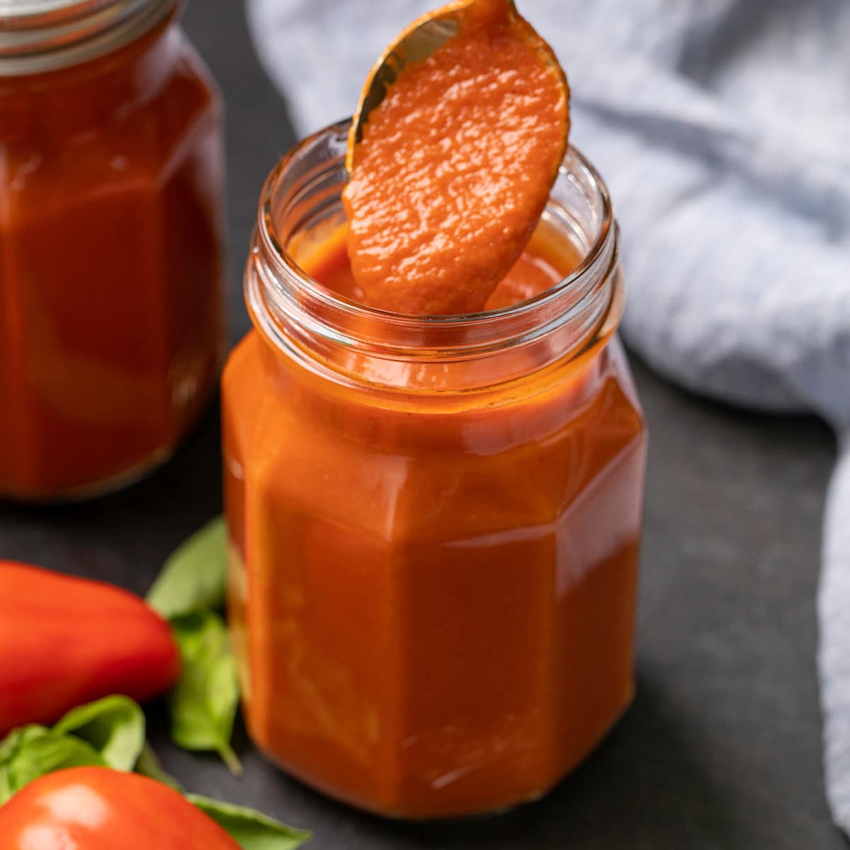 spoonful of gluten free enchilada sauce in glass jar with basil and tomatoes on black surface