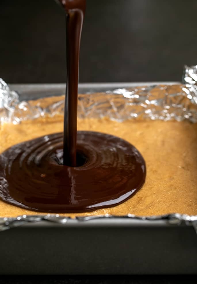 Melted chocolate mixture pouring onto peanut butter bars in square metal pan