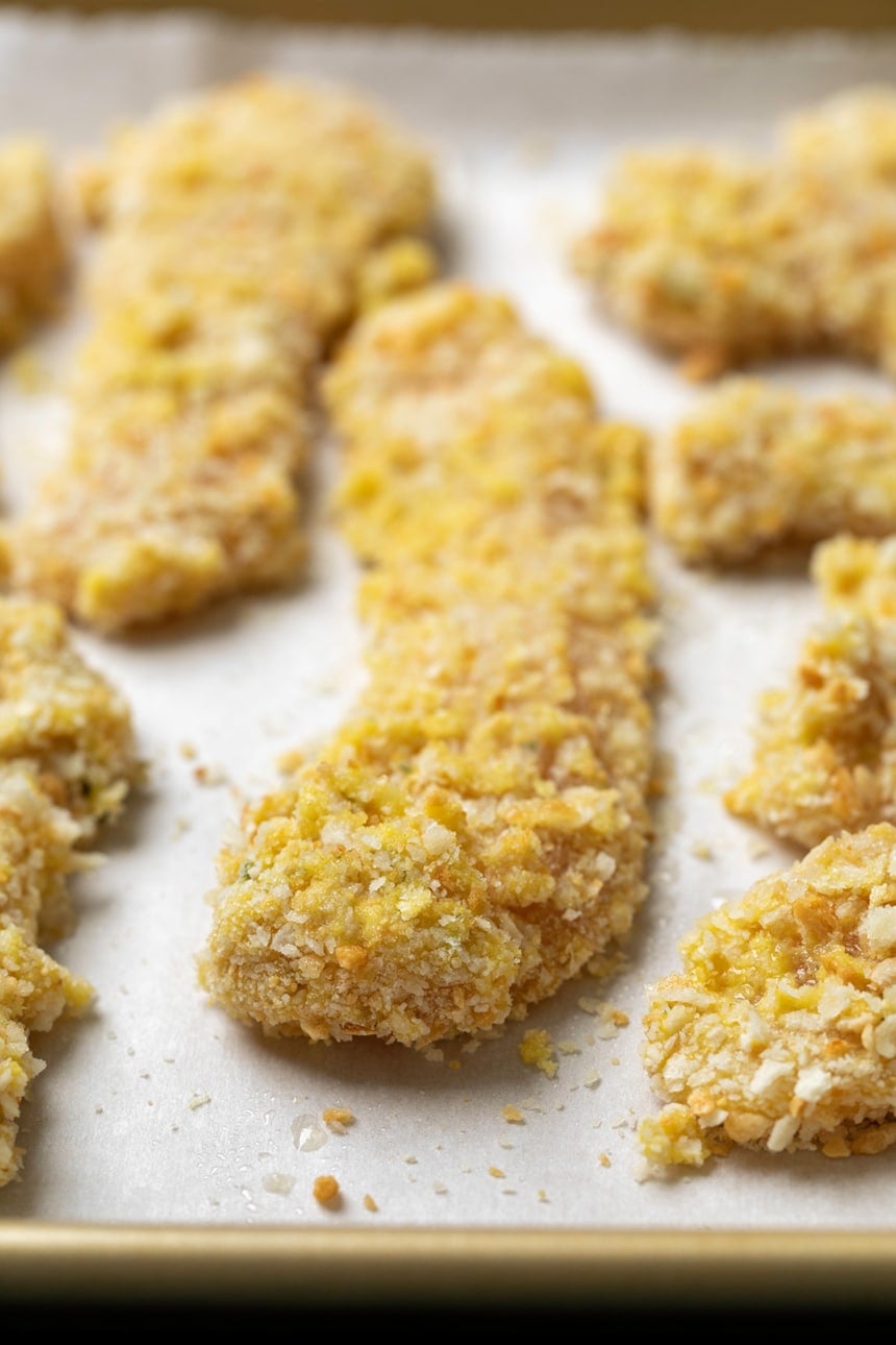 Raw chicken strips coated in breadcrumbs on white paper on gold baking tray