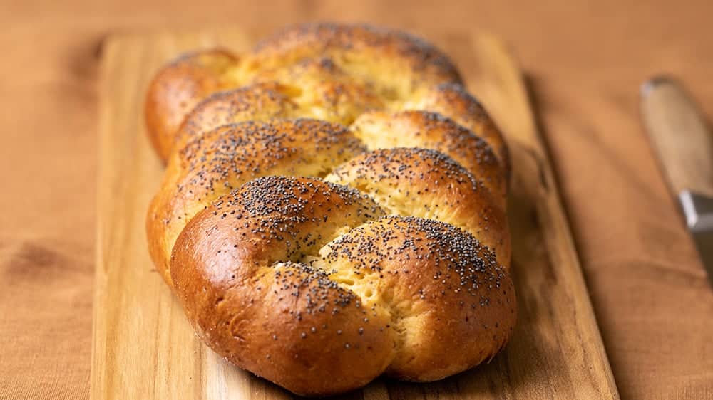 Baked loaf of gluten free challah bread with poppy seeds on wooden cutting board on brown cloth with bread knife