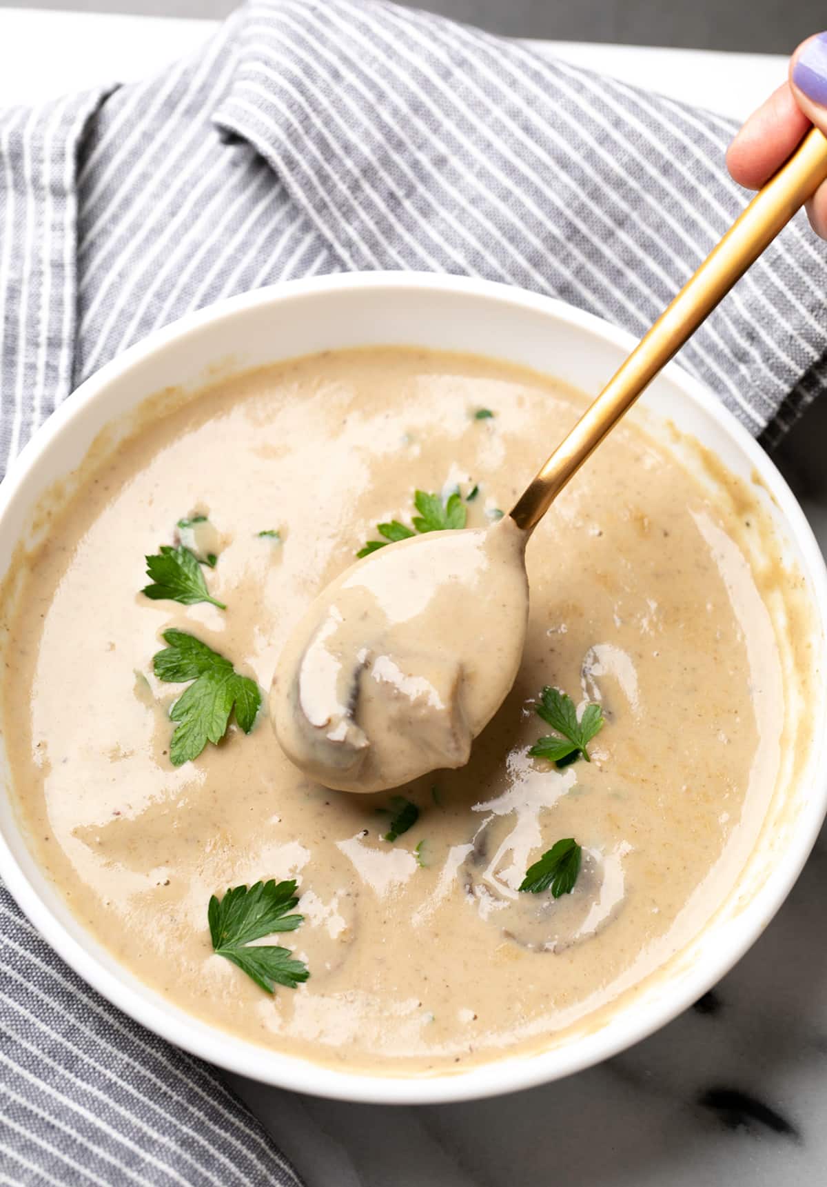 white flat bowl with light brown cream of mushroom soup on gold spoon with green parsley leaves