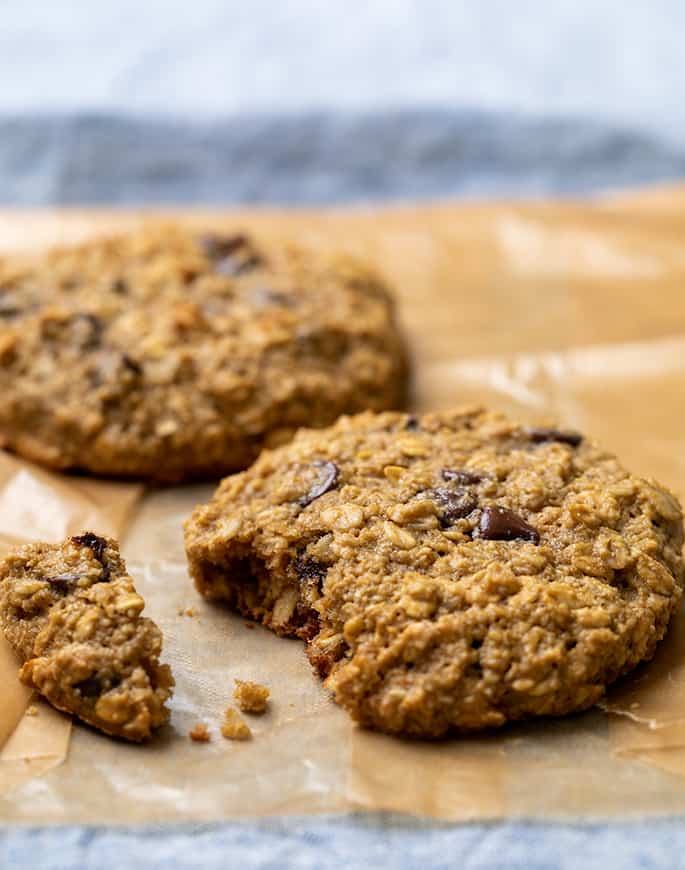 two oatmeal cookies with chocolate chips on brown wax paper, one cookie with a bite removed and crumbs