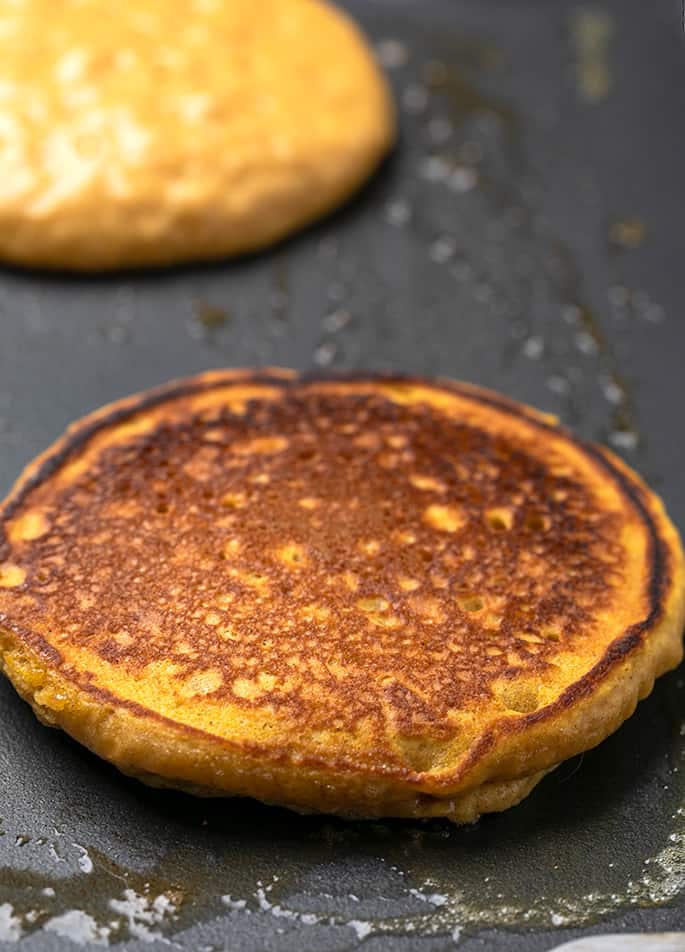 Cooked and browned pumpkin pancake on buttered griddle with one raw on top pancake in background