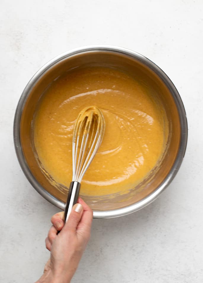 Hand holding metal whisk in orange pancake batter in round metal bowl on white surface