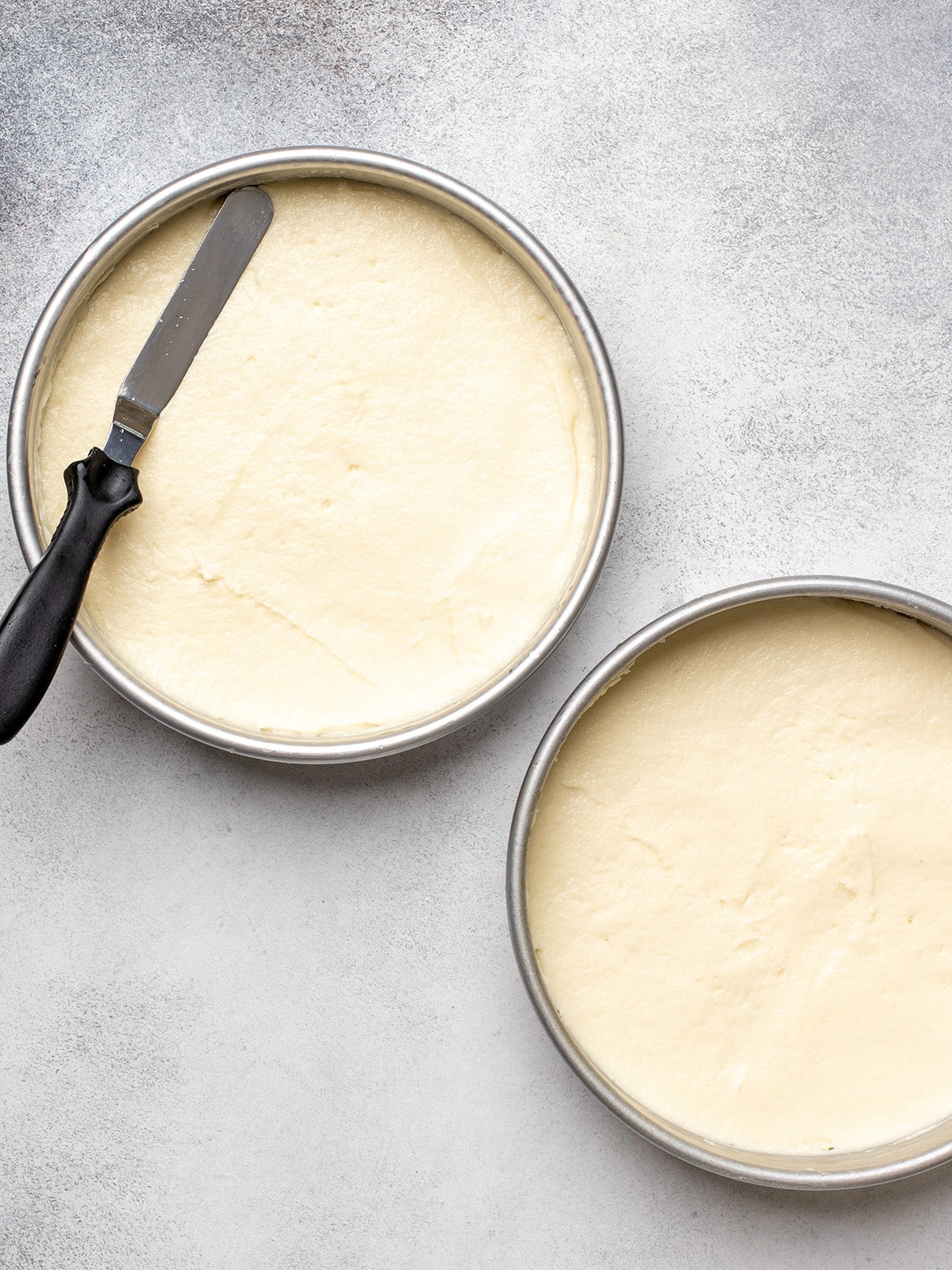 Two round cake pans with gluten free cake batter and an offset spatula on top of one