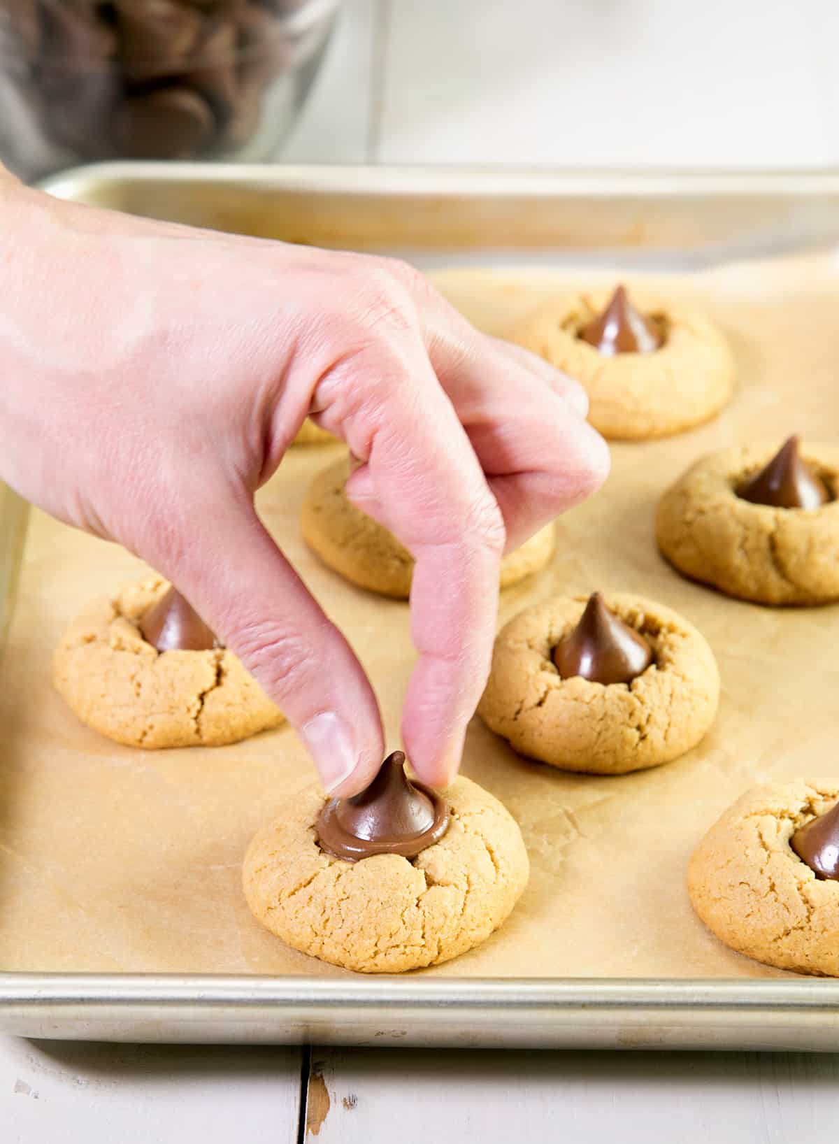 A tray with peanut butter blossom cookies with a hand pressing a chocolate kiss into the top of one just-baked cookie