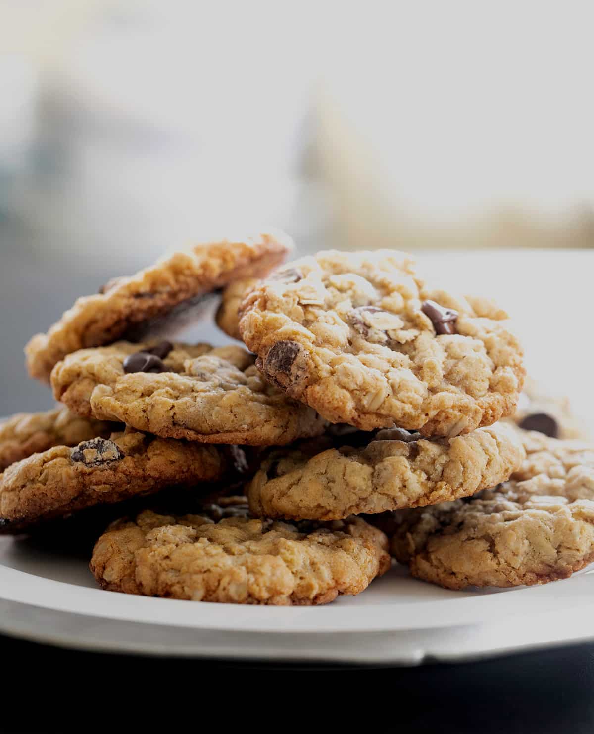A plate of 8 baked cookies with oats and chocolate chips in a haphazard pile on a white serving platter with a metal edge