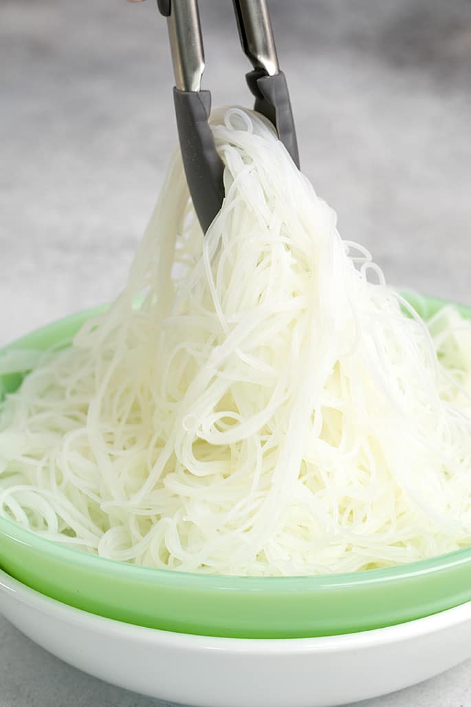 Tongs picking up thin white gluten free rice noodles from green bowl