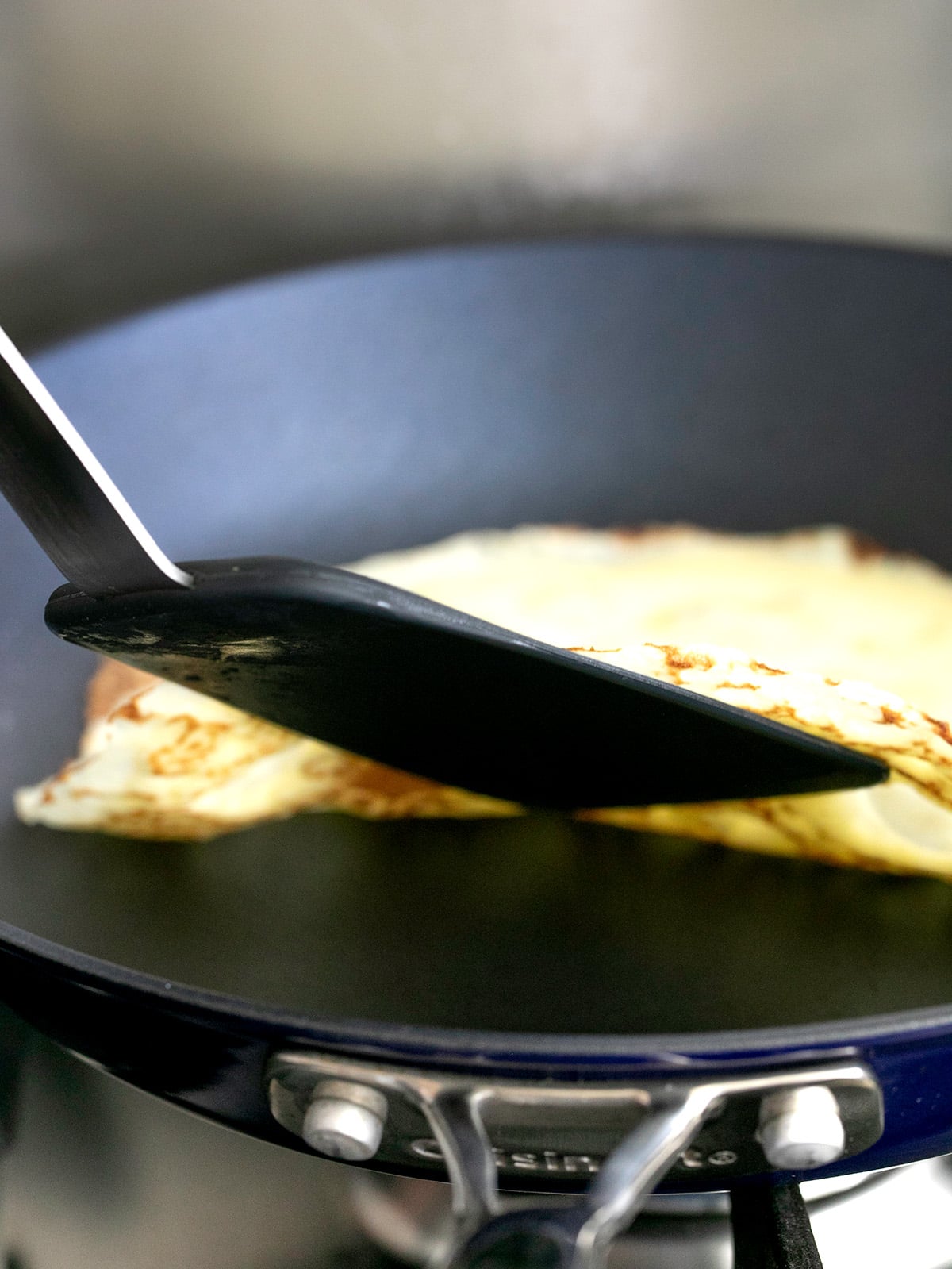 Black spatula lifting edge of gluten free crepes in blue cuisinart nonstick pan
