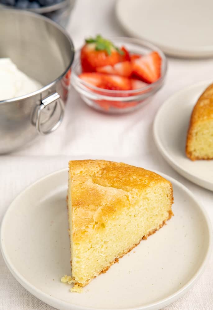 Slice of gluten free olive oil cake on white plate with toppings in background