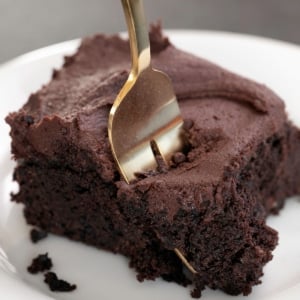 square piece of brown chocolate cake with brown frosting on a small white plate with a gold fork