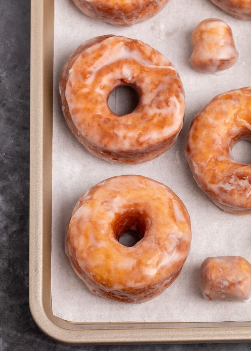 3 brown gluten free donuts with holes with white glaze with 2 glazed donut holes on white paper on gold rimmed baking sheet