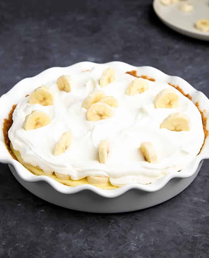 Banana slices in whipping cream on top of more bananas and yellow custard in white pie plate