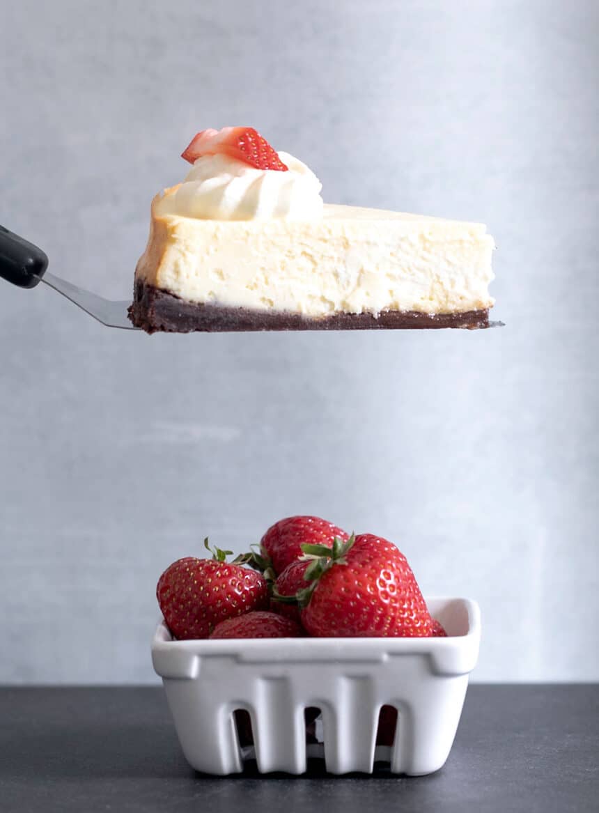 side view of slice of gluten free cheesecake on metal spatula with dollop of whipped cream and strawberry slice over small basket of whole strawberries