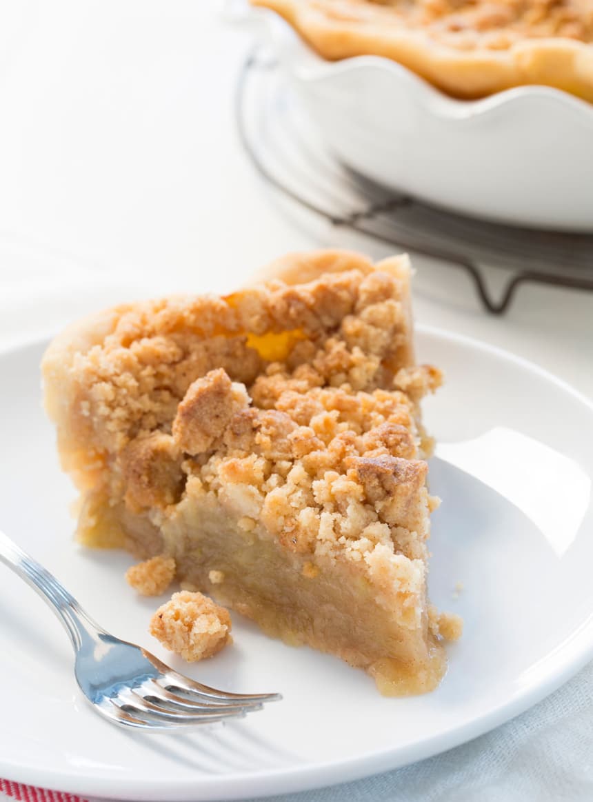 A thick slice of apple crumb pie on a white plate, showing a golden, buttery crumble topping and tender apple filling.