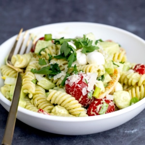 gluten free pasta salad with light tan cooked fusilli, fresh green parsley, red grape tomatoes, white pearl mozzarella cheese and dressing in white bowl with gold fork