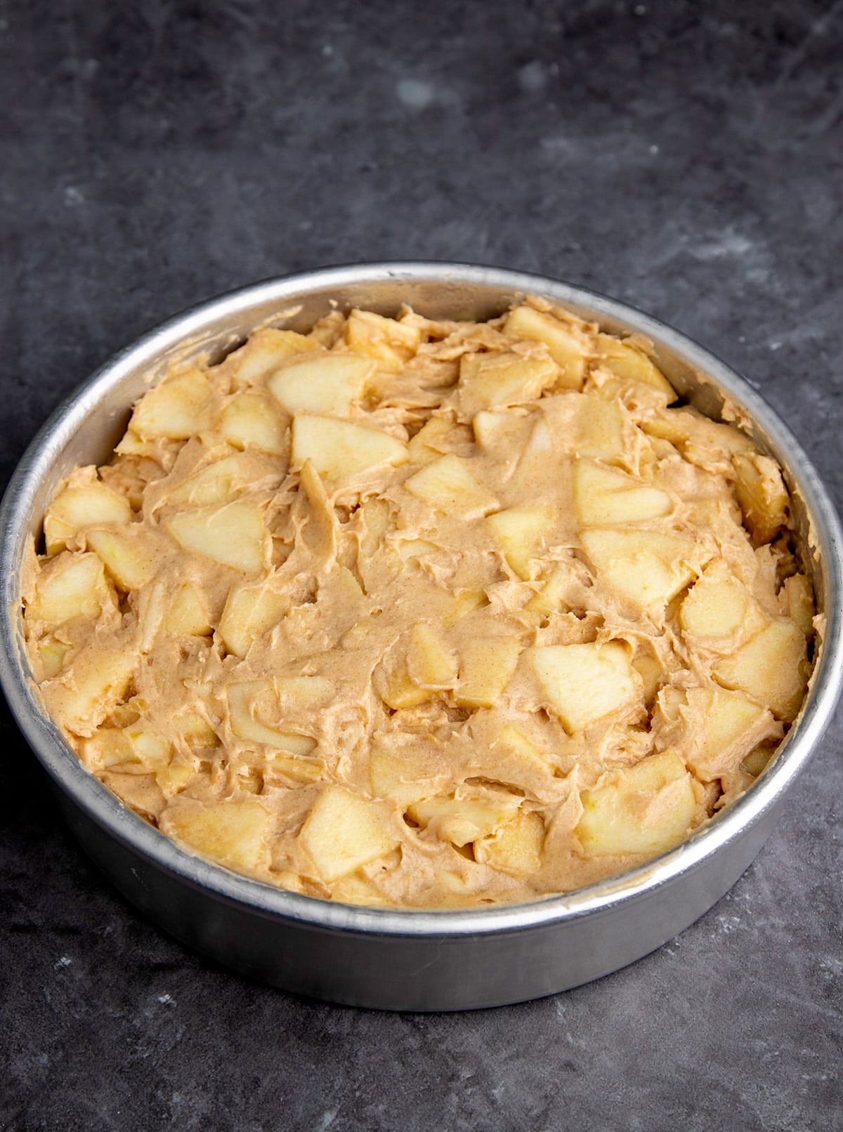 a round cake pan with raw apple cake batter with studded apples mixed throughout on a gray surface