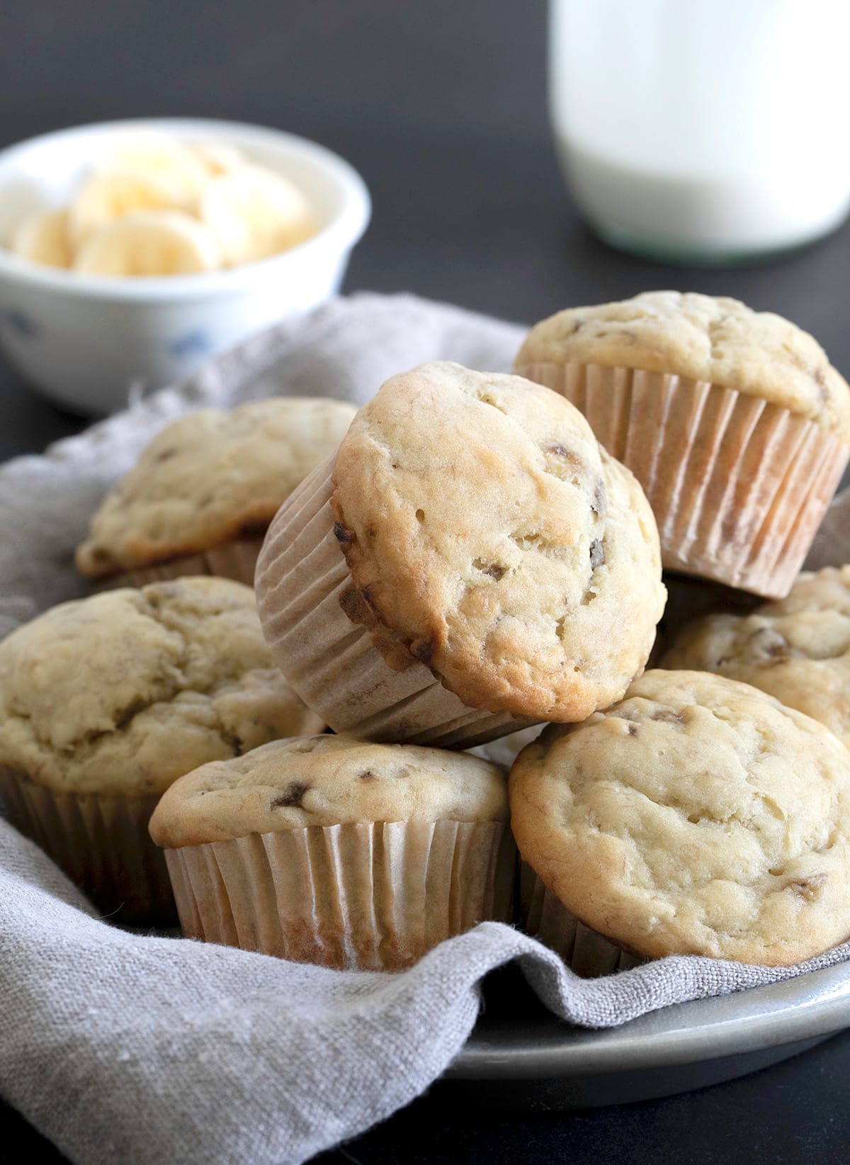 A pile of baked muffins with white muffin liners in a flat bowl lined with light tan cloth.
